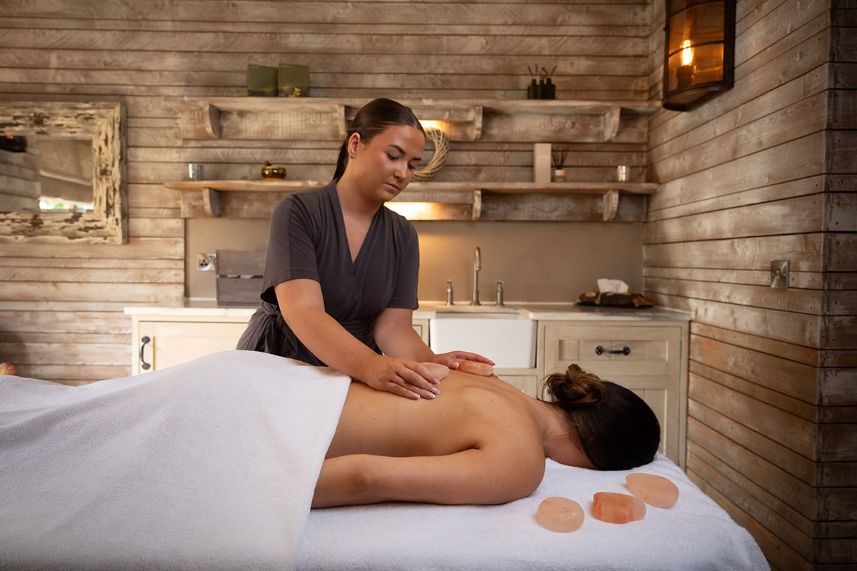 a woman with brown her tied in a loose bun lies face down on towelled table with her upper back exposed and another woman placing pale red smooth stones on her skin