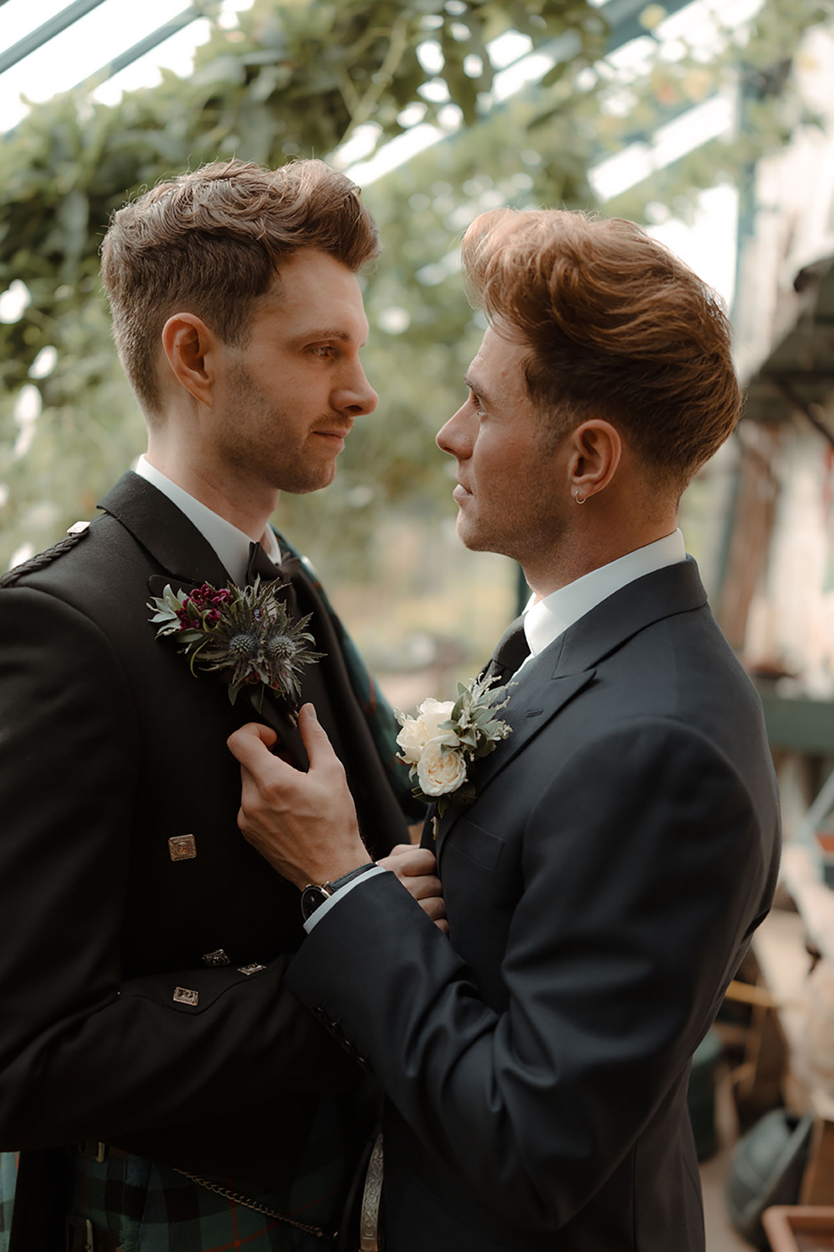 Waist-up portrait of two grooms standing close together, wearing coordinating boutonnieres with a Gunn tartan kilt and a navy wedding suit at Newhall Estate