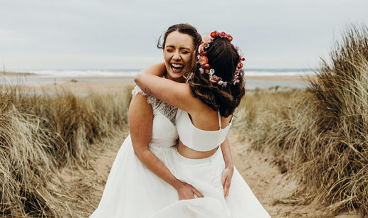 two brides embracing joyfully in a field of tall grass with sand on the ground, one wearing a flower crown