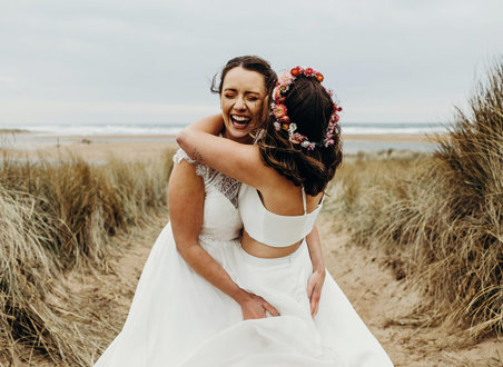 two brides embracing joyfully in a field of tall grass with sand on the ground, one wearing a flower crown