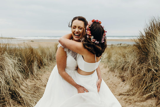 two brides embracing joyfully in a field of tall grass with sand on the ground, one wearing a flower crown