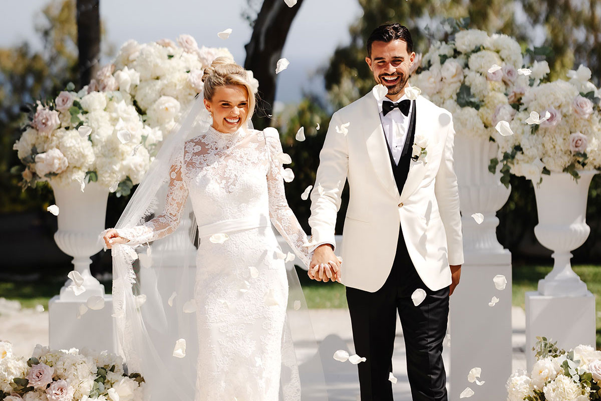 Bride and groom walk hand in hand down the aisle as flower petals fall, surrounded by elegant white and blush floral arrangements. The bride wears a lace long-sleeve wedding dress with a high neck and veil, while the groom wears a white tuxedo jacket with black trousers and bow tie
