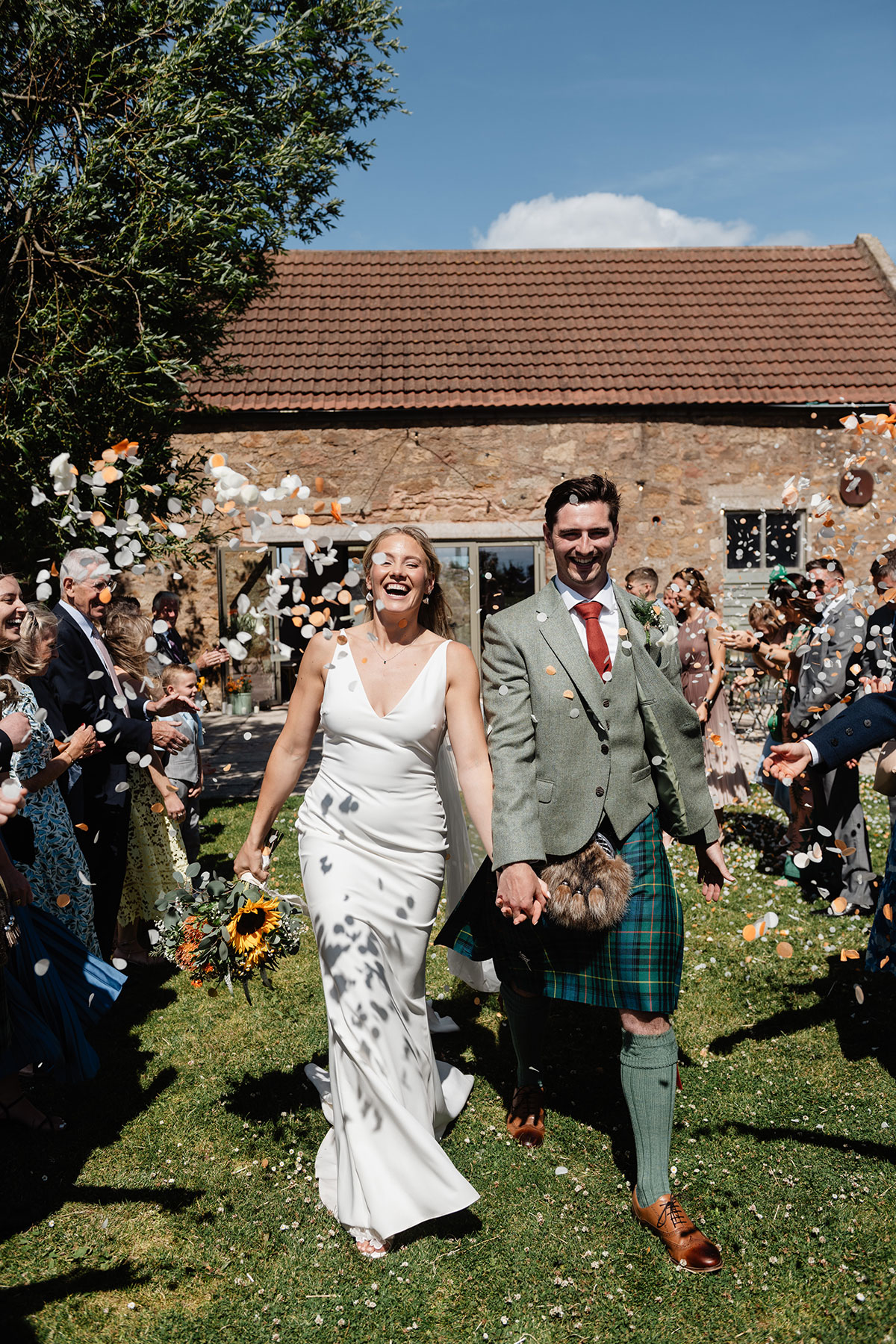 Bride and groom walking through an outdoor confetti toss with guests celebrating under bright summer sunshine