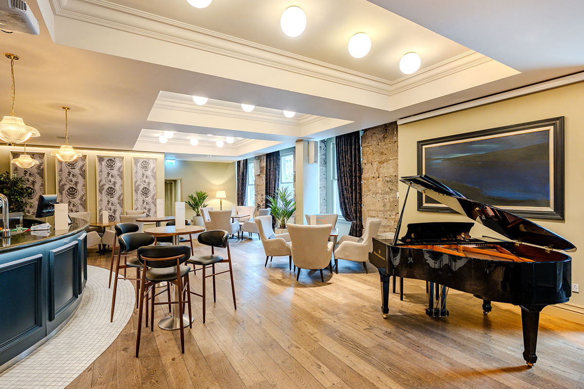  a large room with wooden floors features a bar on one side, a baby grand piano on the other and a mixture of lounge and bar stool seating scattered throughout the room