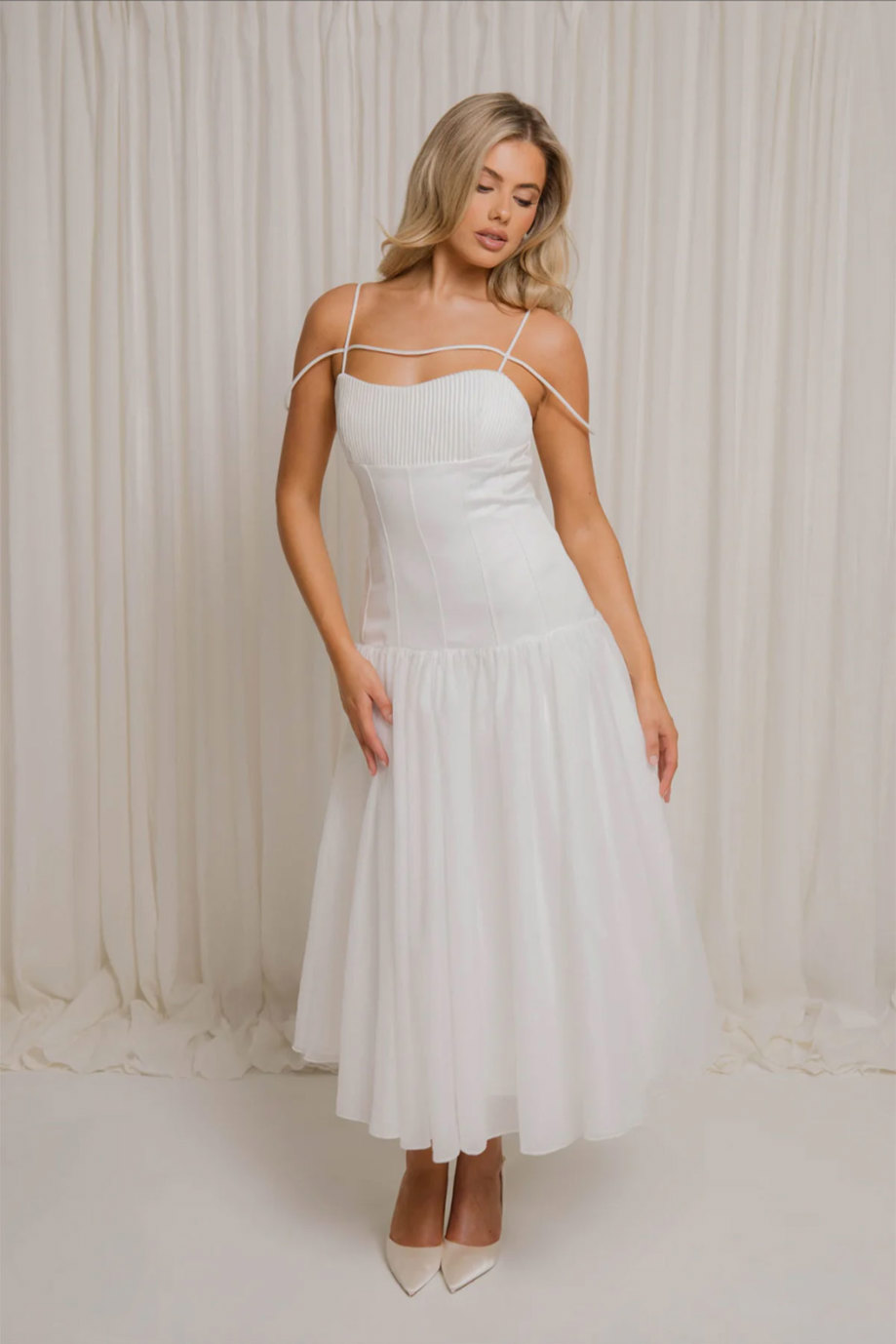blonde model wears white dress with two thin straps and one thin strap across the chest standing in front of cream coloured curtain