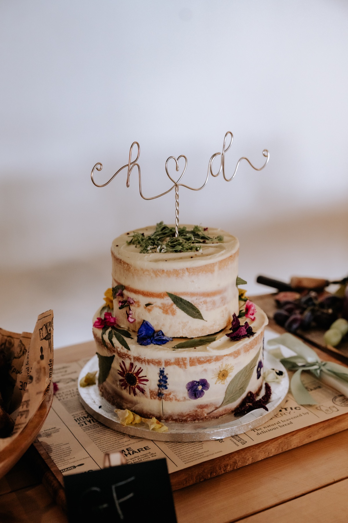Two-tier semi-naked wedding cake decorated with pressed flowers and a Hannah and Donna cake topper.