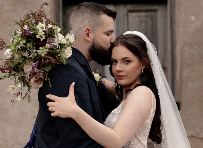 a groom kissing bride on the head. The bride looks serious and straight to camera and she is wearing a veil and holding a large bouquet of flowers