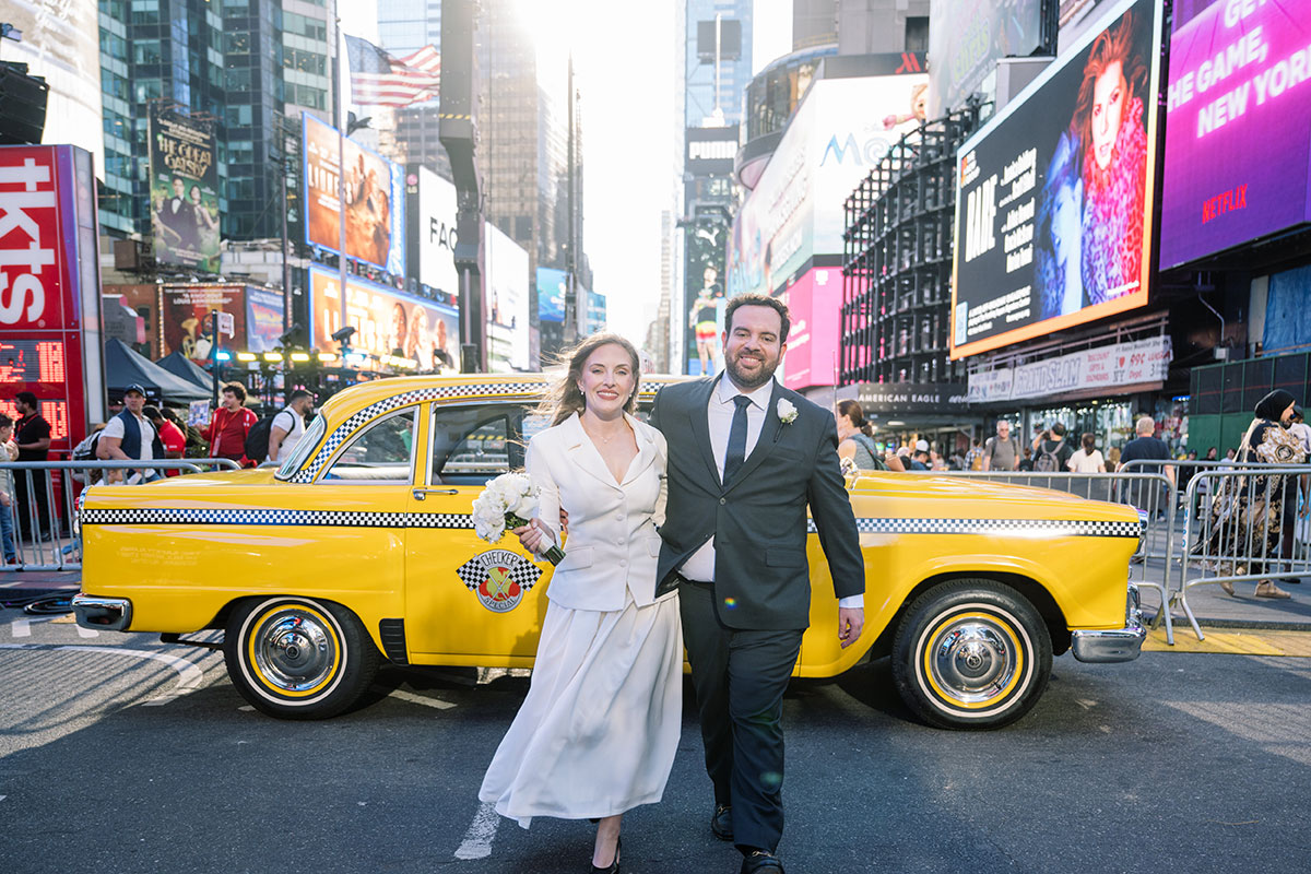 Couple in wedding attire walking through Times Square in front of a yellow taxi with bright billboards and crowds around them