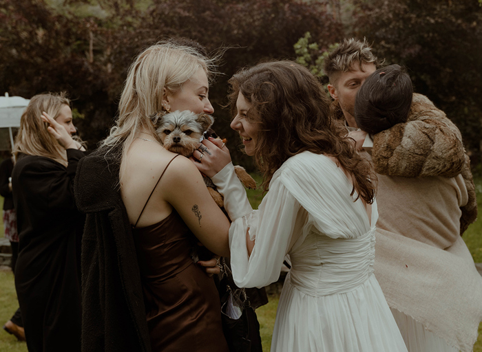 a bride stroking a small dog held in another person's arms. There are other wedding guests in the background