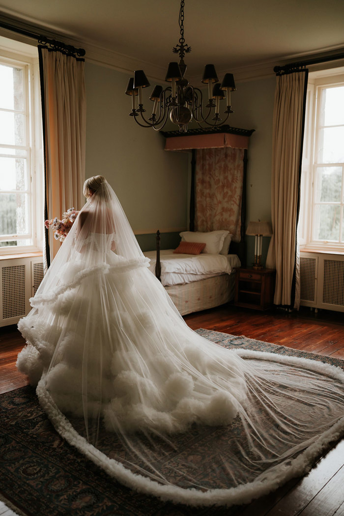 A bride wearing a wedding dress with a large skirt and a long veil facing away from the camera and looking out of a window 