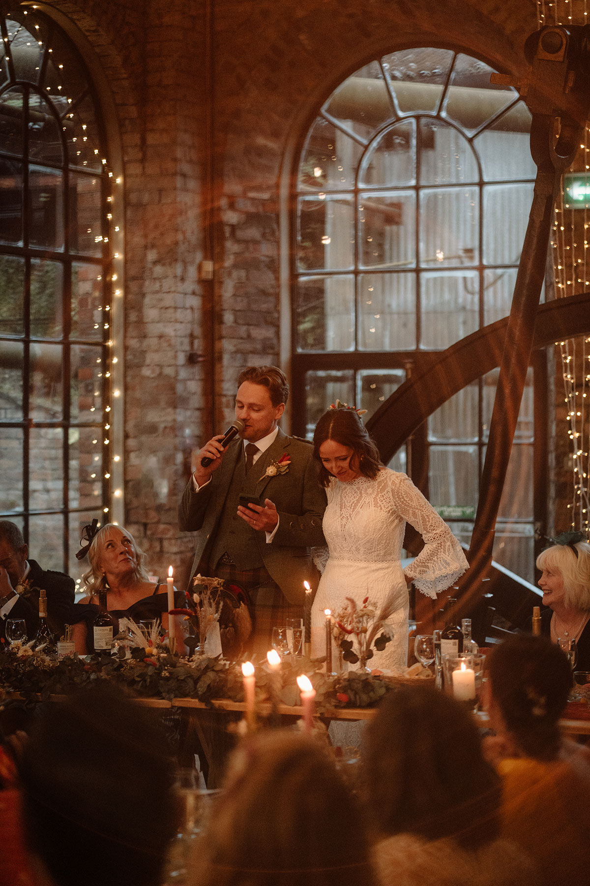 Bride and groom deliver speeches at a candlelit wedding reception inside a restored industrial venue with exposed brick walls and large arched windows
