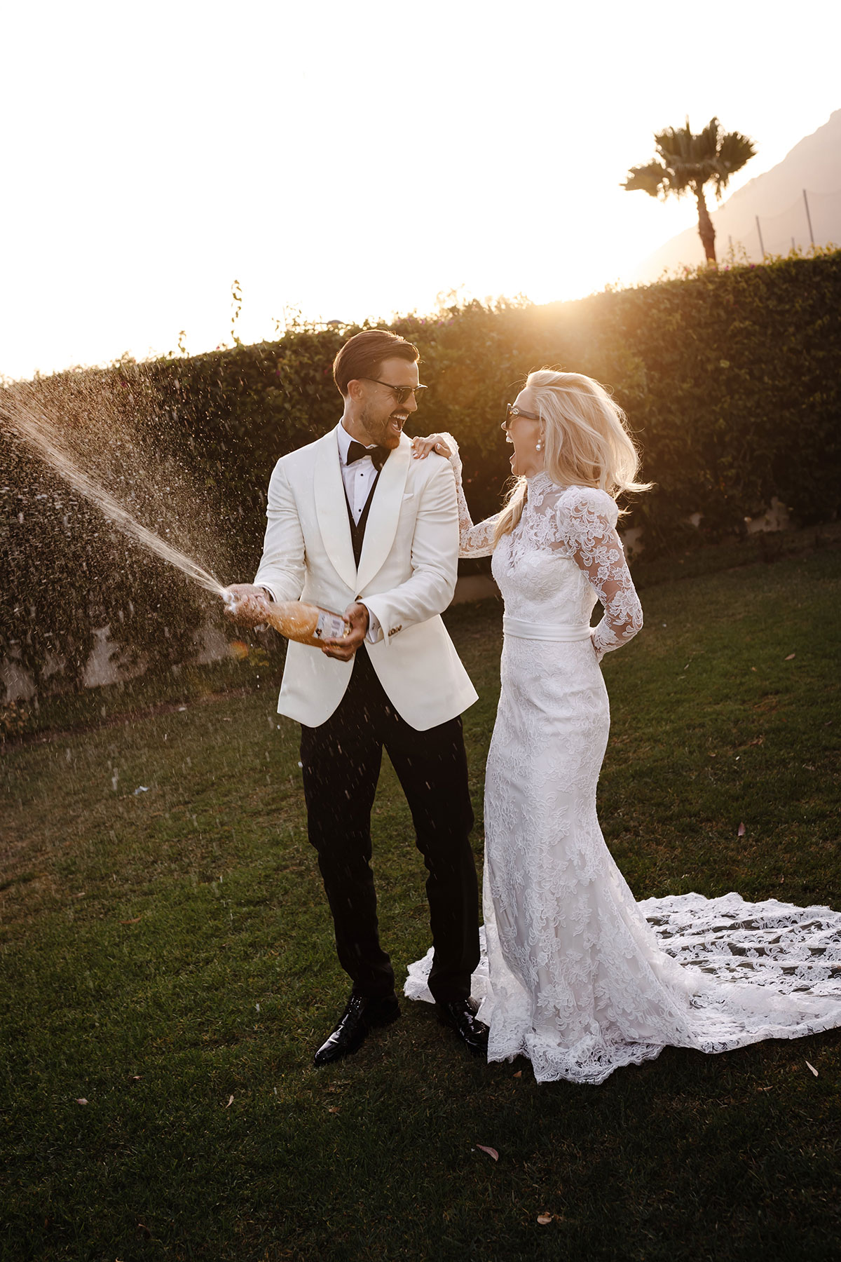 Groom pops champagne as bride laughs beside him in garden at golden hour