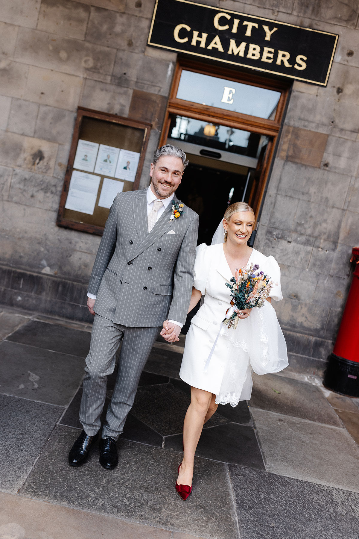 Newlyweds walk out of Edinburgh City Chambers holding hands after their registry office wedding ceremony, smiling and surrounded by historic stone architecture.
