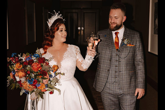 a bride in a white dress holding a large bouquet and groom in a grey suit with an orange tie clink glasses of champagne