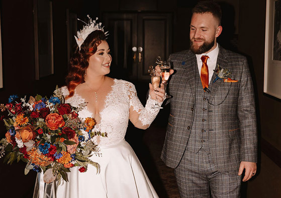 a bride in a white dress holding a large bouquet and groom in a grey suit with an orange tie clink glasses of champagne