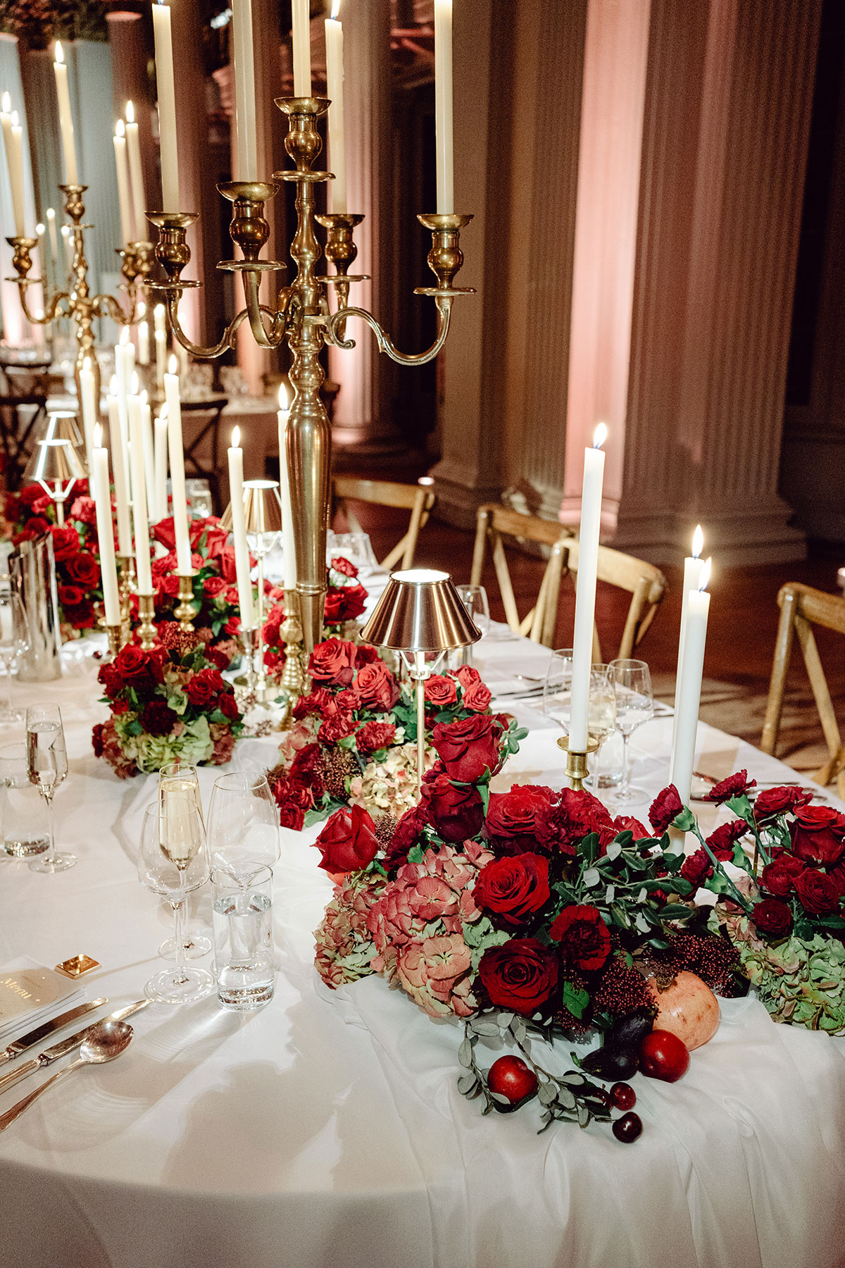 Close-up of red and blush floral arrangements with tall taper candles decorating a long reception table at the Signet Library
