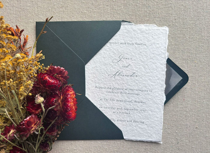 A wedding invitation half in a dark green envelope with red and yellow flowers to the left side
