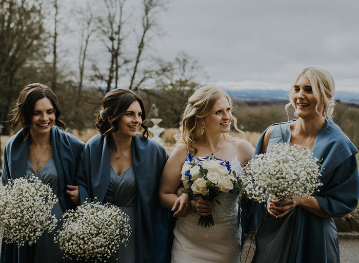 Three women in blue dresses and shawls holding baby's breath flowers and a woman in a white dress holding a bouquet of white roses and purple flowers