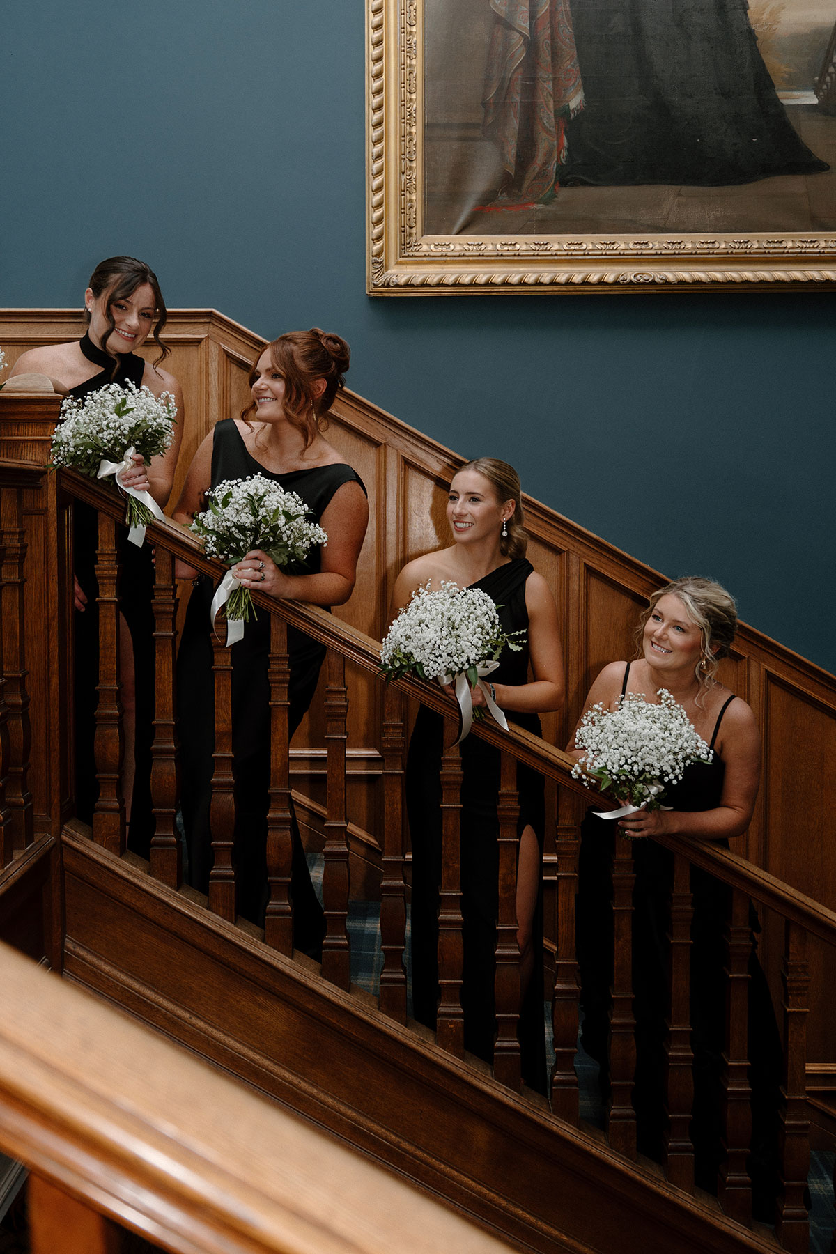 Bridesmaids in black dresses holding white bouquets while posing on a grand wooden staircase inside a Scottish wedding venue
