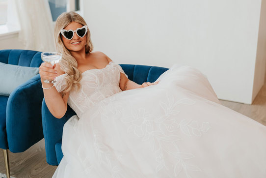 bride sitting in wedding gown on blue couch with pearl sunglasses and a coupe of prosecco