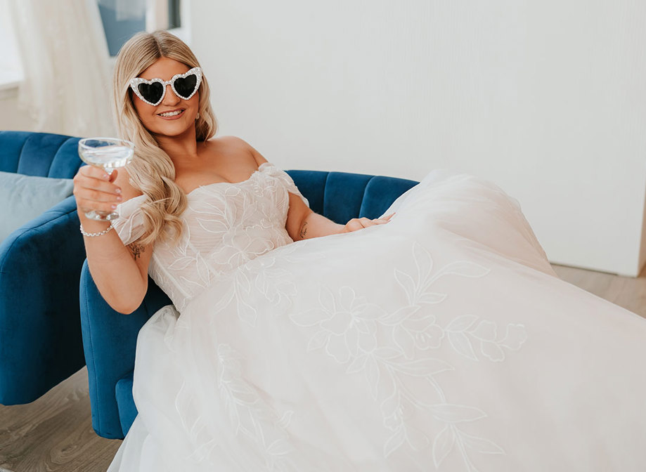 bride sitting in wedding gown on blue couch with pearl sunglasses and a coupe of prosecco