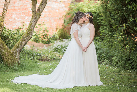 two brides wearing long and fluid wedding dresses standing romantically head-to-head in a walled garden setting at Byre at Inchyra