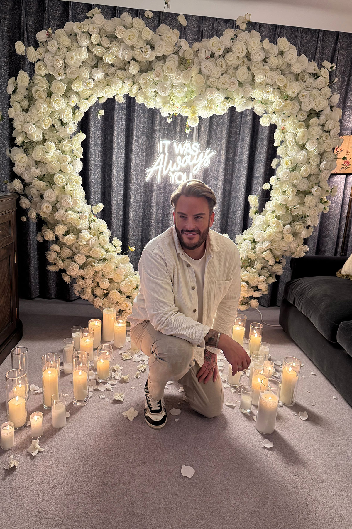 Romantic proposal setup with white rose arch, candles and neon “It was always you” sign, groom kneeling in centre