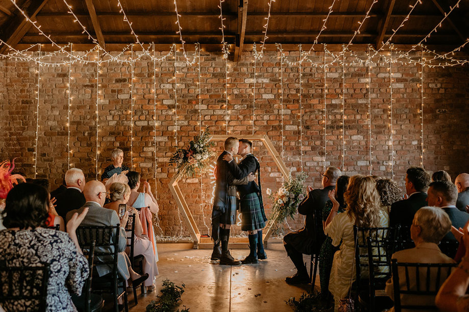 Two Grooms Kissing With Red Brick Wall In Background And Guests Looking On
