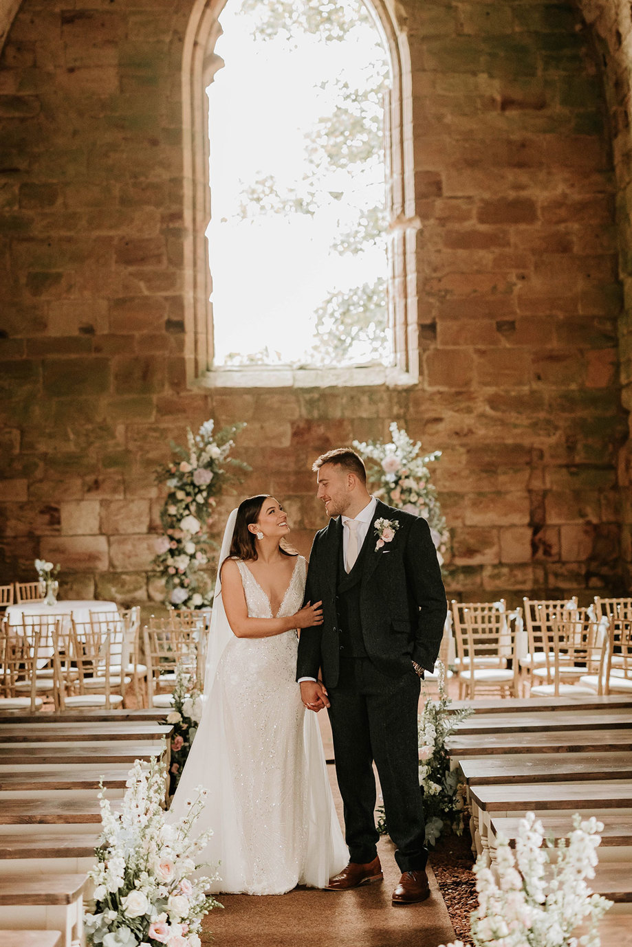 a smiling bride and groom in ruined Dunglass Collegiate Church with floral arrangements on ground