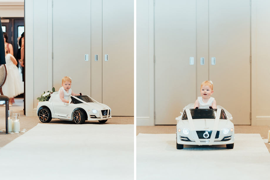 a toddler wearing a white dress sitting on a miniature white car that's travelling on a white carpet 