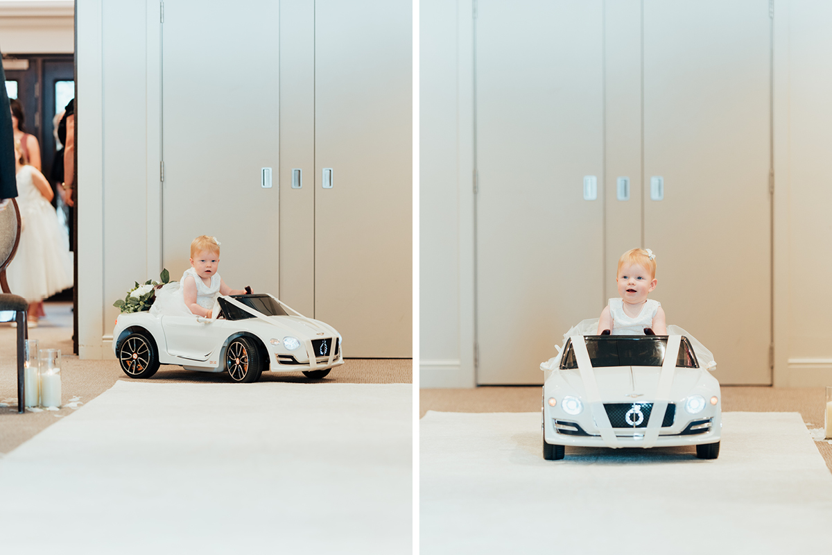 a toddler wearing a white dress sitting on a miniature white car that's travelling on a white carpet