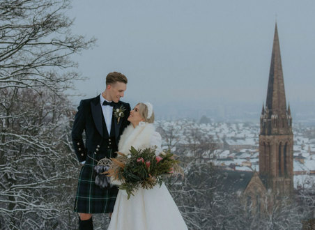 A bride wearing a white dress and white fur wrap standing with a groom wearing a dark kilt in a park in the snow