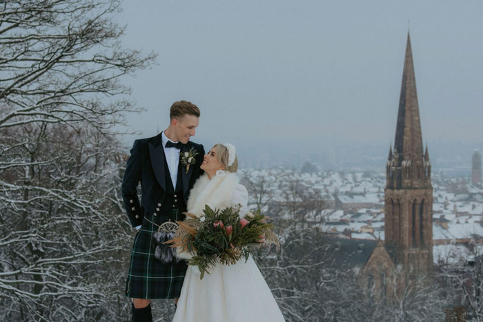 A bride wearing a white dress and white fur wrap standing with a groom wearing a dark kilt in a park in the snow