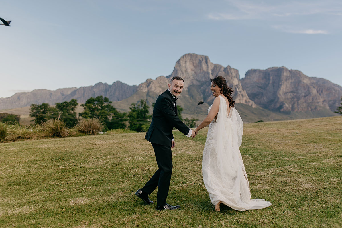 Bride and groom holding hands across the lawns at Boschendal Wine Estate with Cape Town mountain views behind them