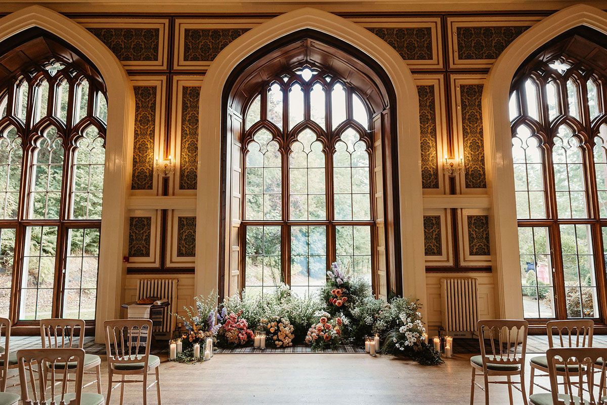 Grand ceremony setup in a historic hall with tall arched windows, ornate wall detailing, floral arrangements, and candles lining the floor