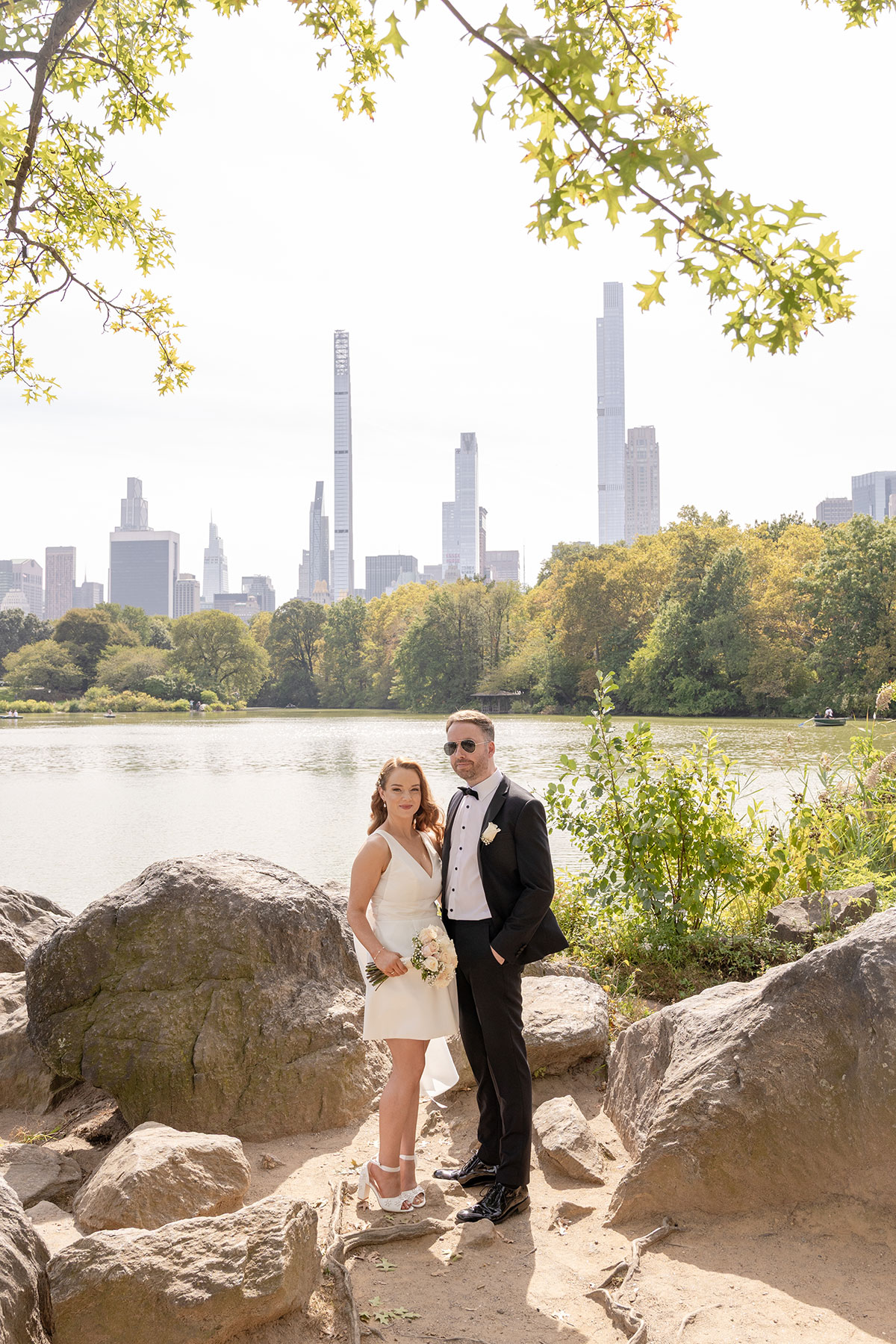 Couple in wedding attire standing on rocks beside a lake in Central Park with New York skyline in the background and leafy branches framing the scene