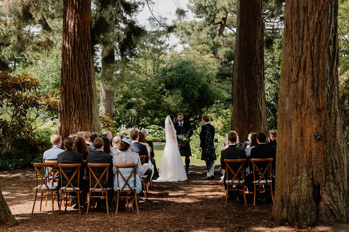 bride and groom stand at their wedding ceremony, outdoors in between tall trees, with guests seated either side of them on wooden chairs