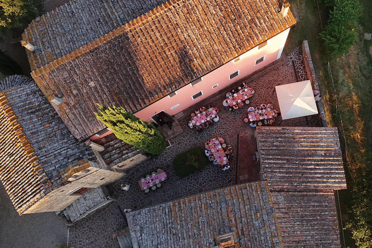 Aerial view of a rustic Tuscan villa courtyard with terracotta roofs and long dining tables set outdoors with pink linens for a wedding reception