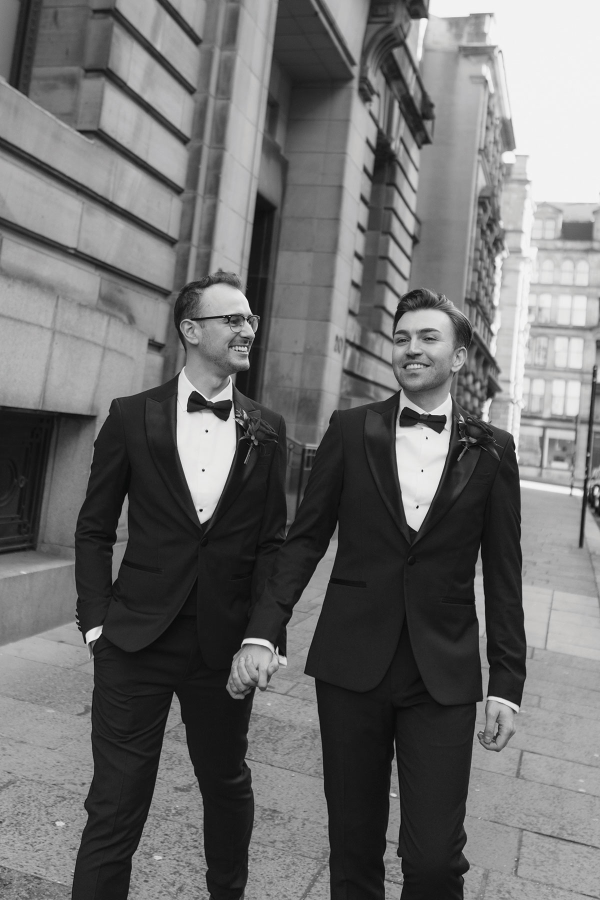 Two grooms in black tuxedos walking hand in hand through Glasgow city centre during their wedding portrait session.