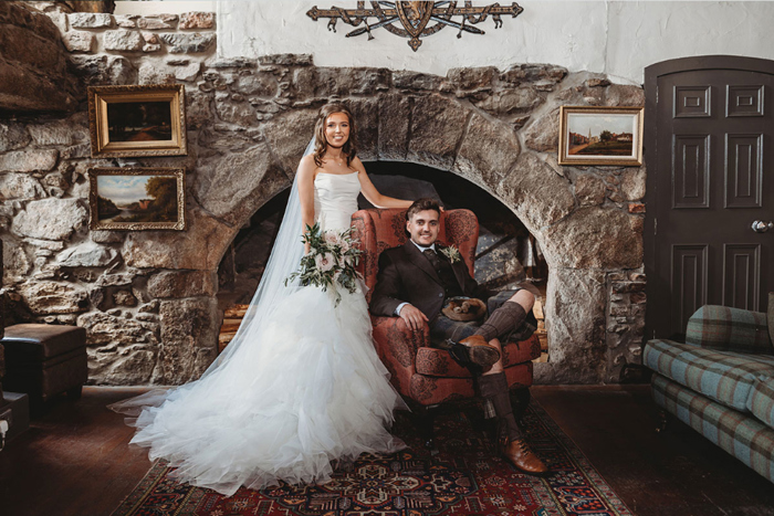 Couple portrait with groom sitting on chair and bride standing against stone wall backdrop