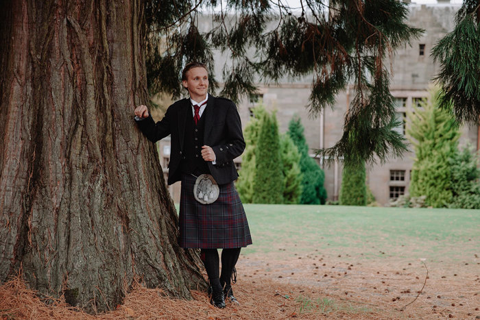 blonde man in full kilt outfit, sporran and red tie, leans against a large tree with heaps of pine needles on the ground around his feet