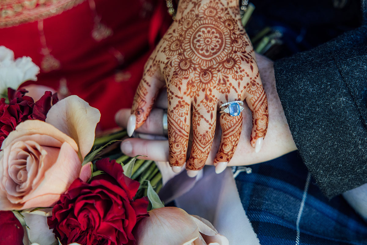 Close-up of the bride's engagement ring and intricate henna as she holds hands with her groom, surrounded by red and blush wedding flowers