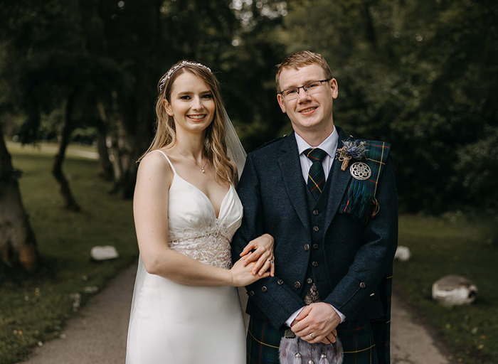 A bride in a white dress and a groom in a dark blue kilt link arms and smile at the camera