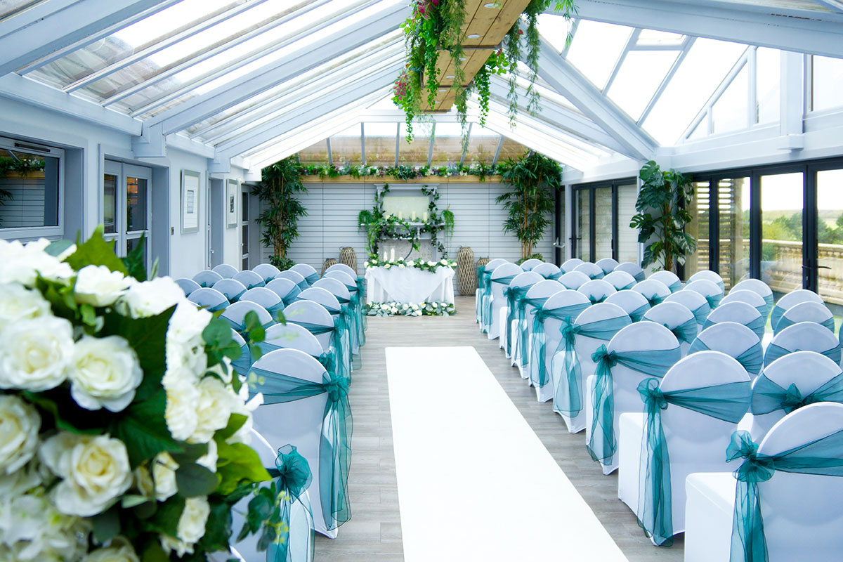 Rooftop Conservatory at The Gailes Hotel & Spa arranged for a wedding ceremony with white chairs, teal sashes and greenery throughout.
