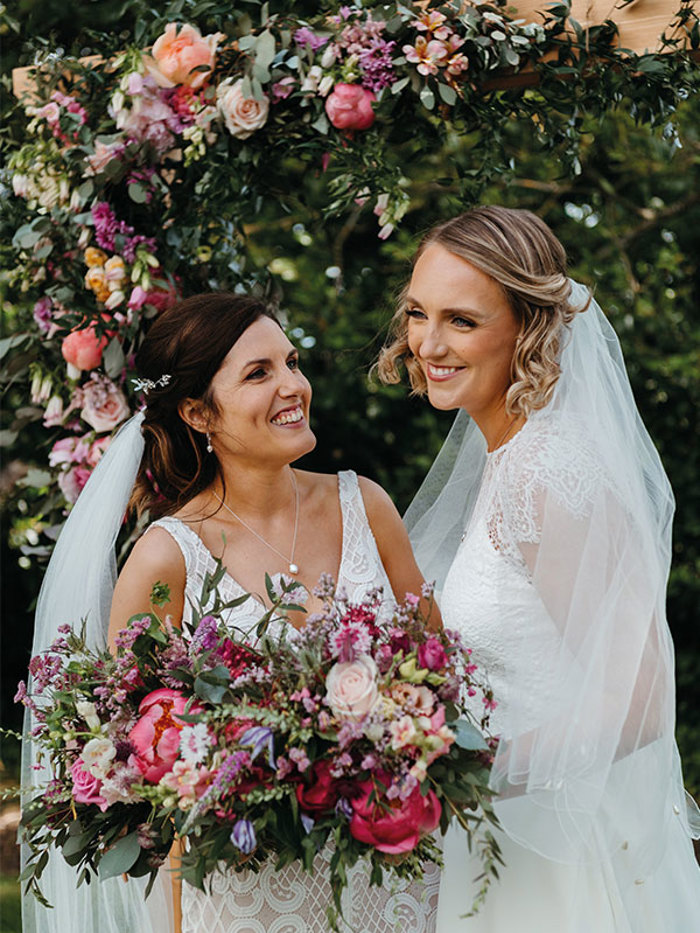 Two brides smiling and holding a bouquet with pink peonies and other pink and purple flowers