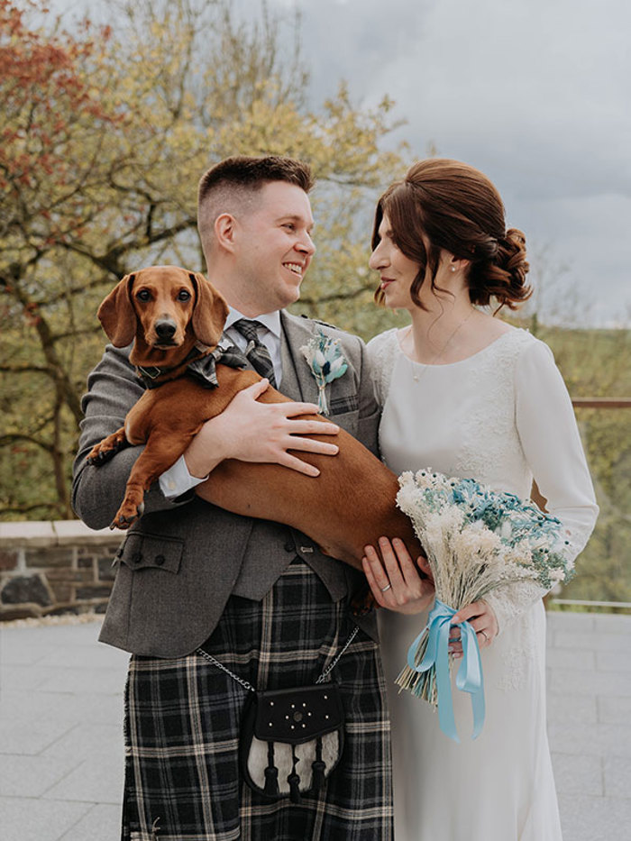 A man in a kilt holds a brown sausage dog who is wearing a bowtie as he stands next to a woman in a white dress