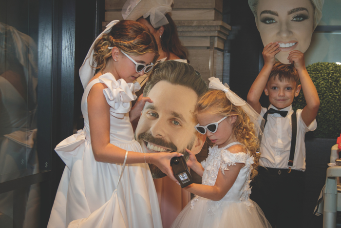 two young girls in white dresses and sunglasses and young boy in bow tie and braces holding large cut out heads of bride and groom