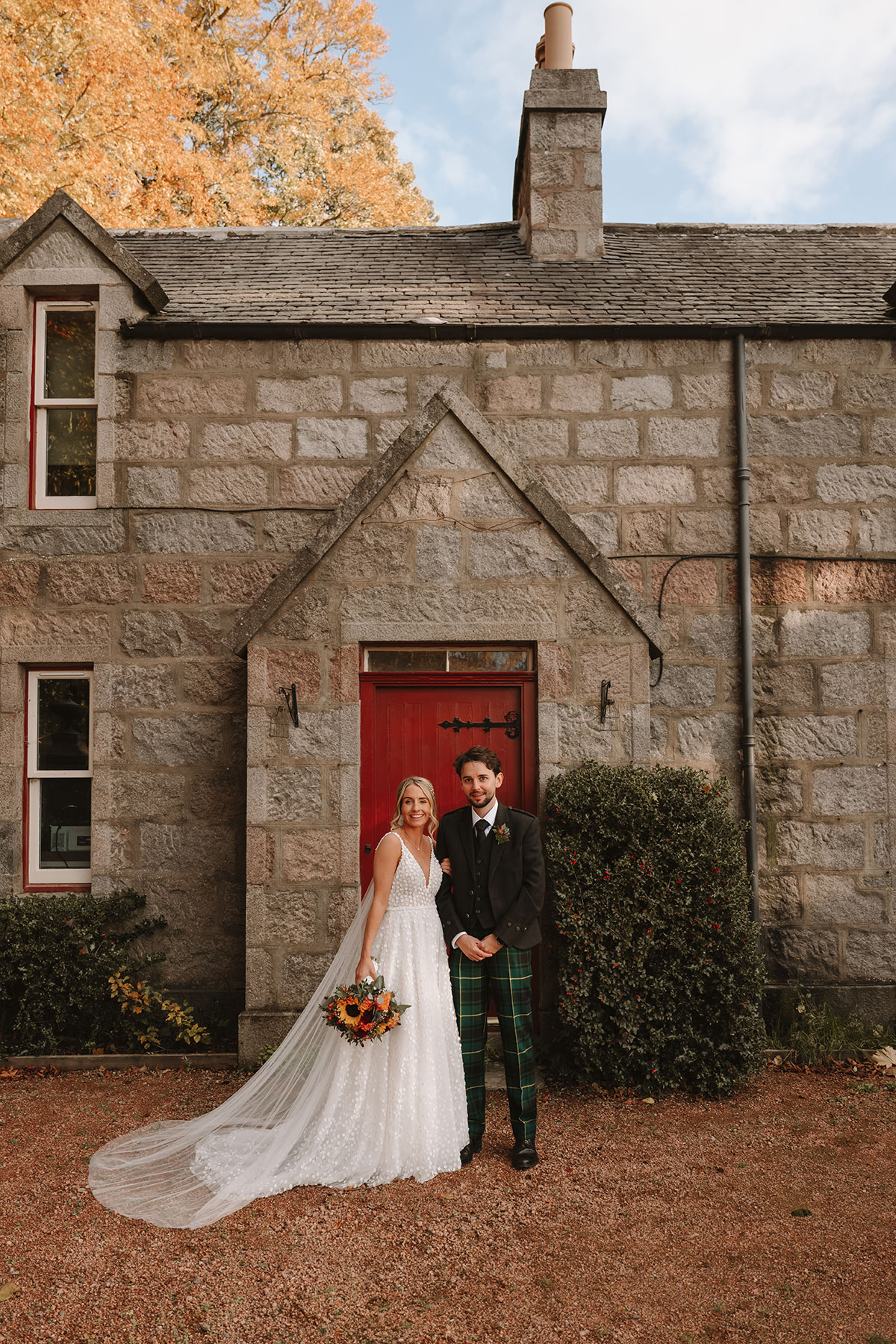 Bride and groom standing outside a brick cottage with a red door, smiling at the camera