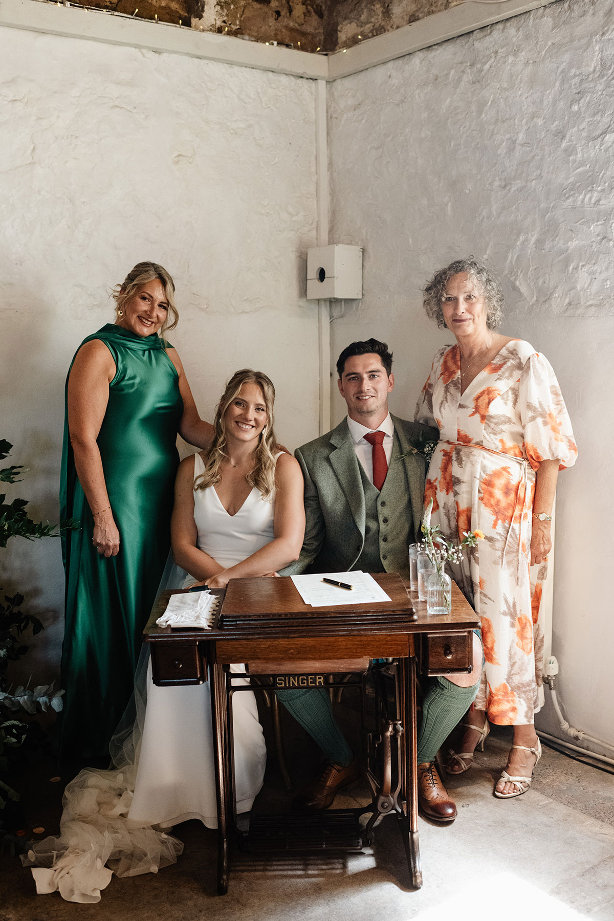 Bride and groom seated at a vintage Singer sewing table signing the marriage register, with two women standing beside them