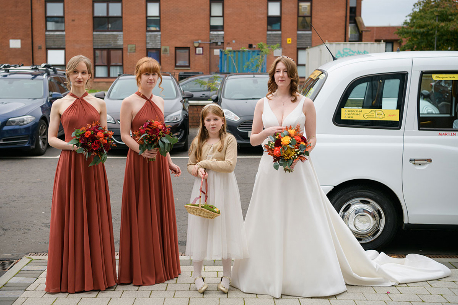 Bride smiles beside flower girl wearing white dress and bridesmaids wearing rust-coloured dresses